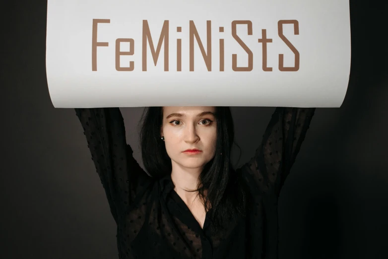 a woman holding a sign that says feminists, an album cover, by Anna Füssli, looking straight to camera, sheryl sandberg, mantis, center view
