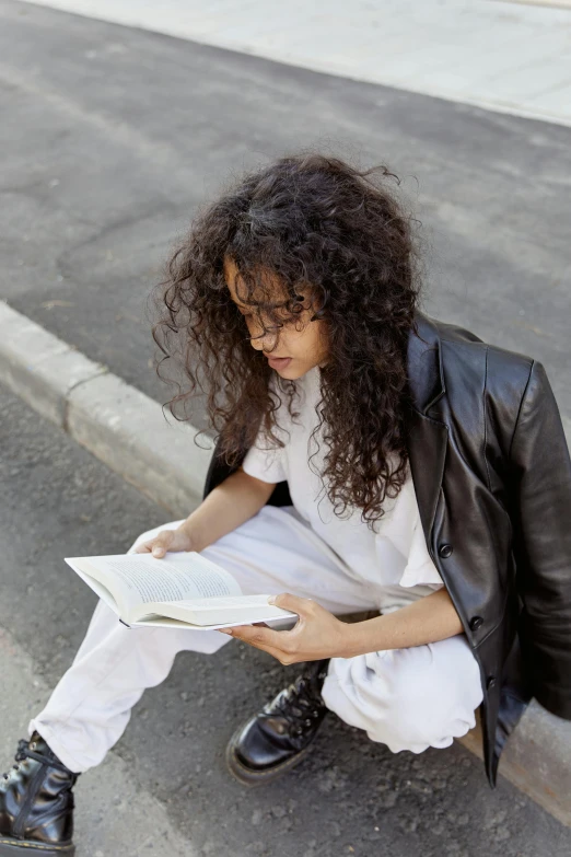 a woman sitting on a curb reading a book, long wild black curly hair, white and black clothing, androgynous person, wearing off - white style