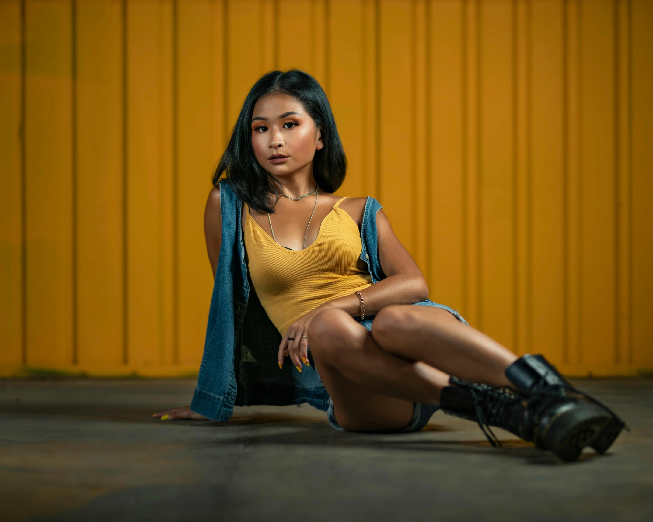 a woman sitting on the ground in front of a yellow wall, by Ric Estrada, pexels contest winner, hyperrealism, wearing a camisole and boots, young asian woman, badass pose, looking confident