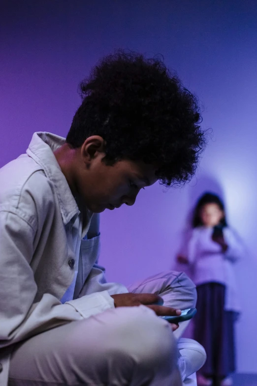 a man sitting on a stool looking at a cell phone, by artist, trending on unsplash, interactive art, purple scene lighting, black teenage boy, teenage girl, in a gallery setting