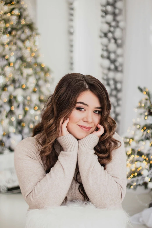 a woman sitting in front of a christmas tree, by Julia Pishtar, trending on pexels, brown wavy hair, chloe bennet, pastel', winking at the camera
