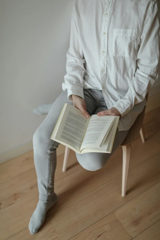 a man sitting on a chair reading a book, by Grytė Pintukaitė, pexels contest winner, cream colored peasant shirt, minimal clothing, light grey, wearing pants