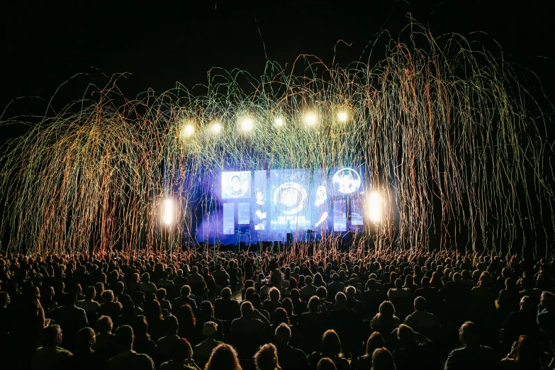 a large group of people standing in front of a stage, by Gavin Nolan, pexels contest winner, kinetic art, neurons firing, fireworks in background, huge viewscreen at front, joel fletcher