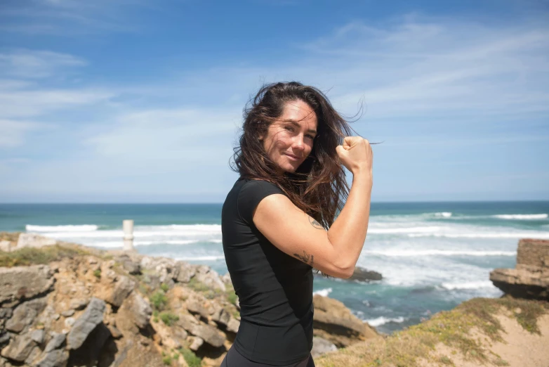 a woman standing on top of a cliff next to the ocean, a portrait, showing off biceps, windy mane, avatar image, manuka