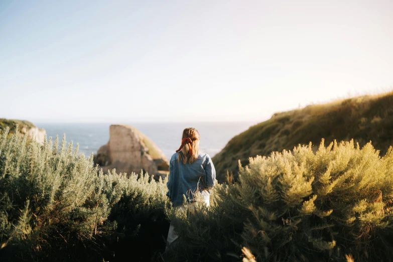 a woman standing on top of a lush green hillside, by Jessie Algie, pexels contest winner, happening, coastal cliffs, denim, back lit, manuka