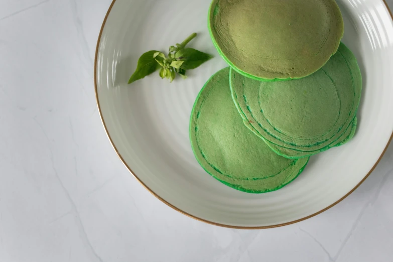 a white plate topped with green pancakes on top of a table, a pastel, inspired by Art Green, unsplash, process art, steamed buns, aerial view. hand stitching, glossy surface, natural soft rim light