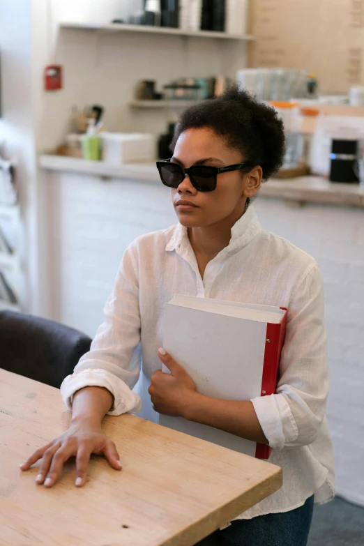 a woman sitting at a table with a book, wearing sunglasses, schomburg, holding notebook, wearing a linen shirt