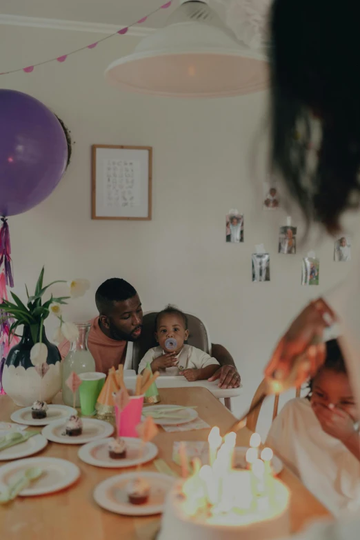 a woman blowing out candles on a birthday cake, a colorized photo, pexels contest winner, father with child, at a dinner table, essence, party balloons