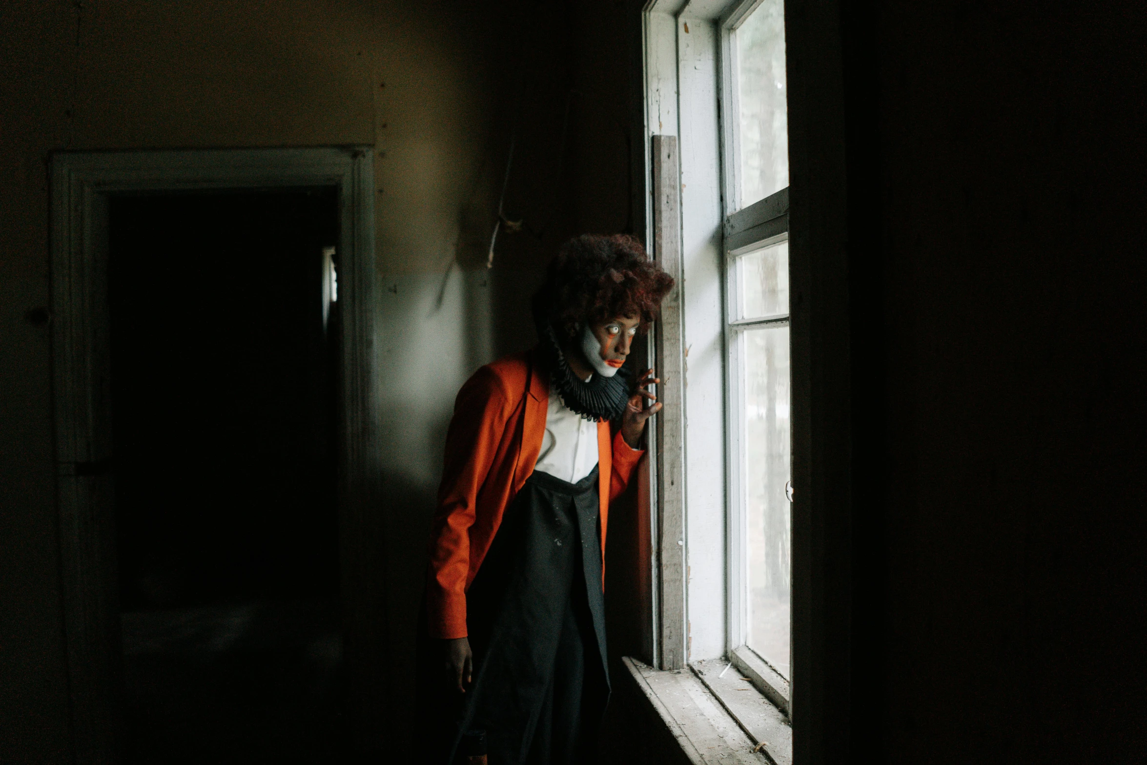 a woman standing by a window in a dark room, inspired by Elsa Bleda, pexels contest winner, black and orange coat, in a desolate abandoned house, african american woman, androgynous person