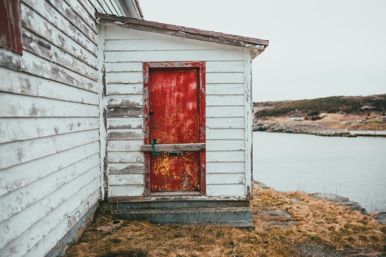 a white house with a red door next to a body of water, by Jessie Algie, pexels contest winner, portrait of a slightly rusty, rugged details, inuit heritage, payne's grey and venetian red