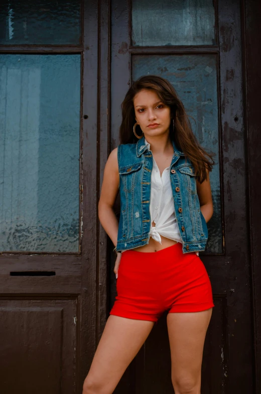 a woman standing in front of a wooden door, inspired by Elsa Bleda, pexels contest winner, wearing red shorts, wearing a jeans jackets, brunette woman, model is wearing techtical vest