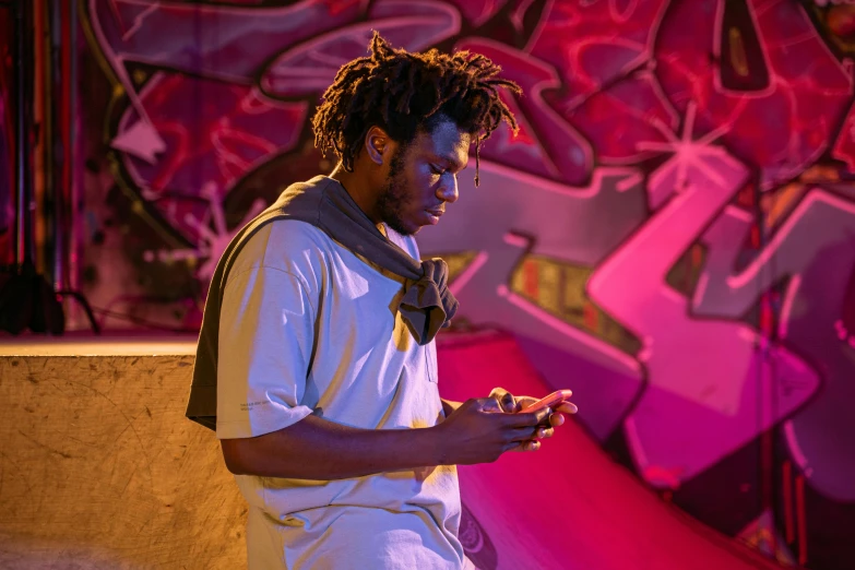 a man standing in front of a wall with graffiti on it, trending on pexels, process art, 2 rappers on stage at concert, looking at his phone, dreads, evening lighting
