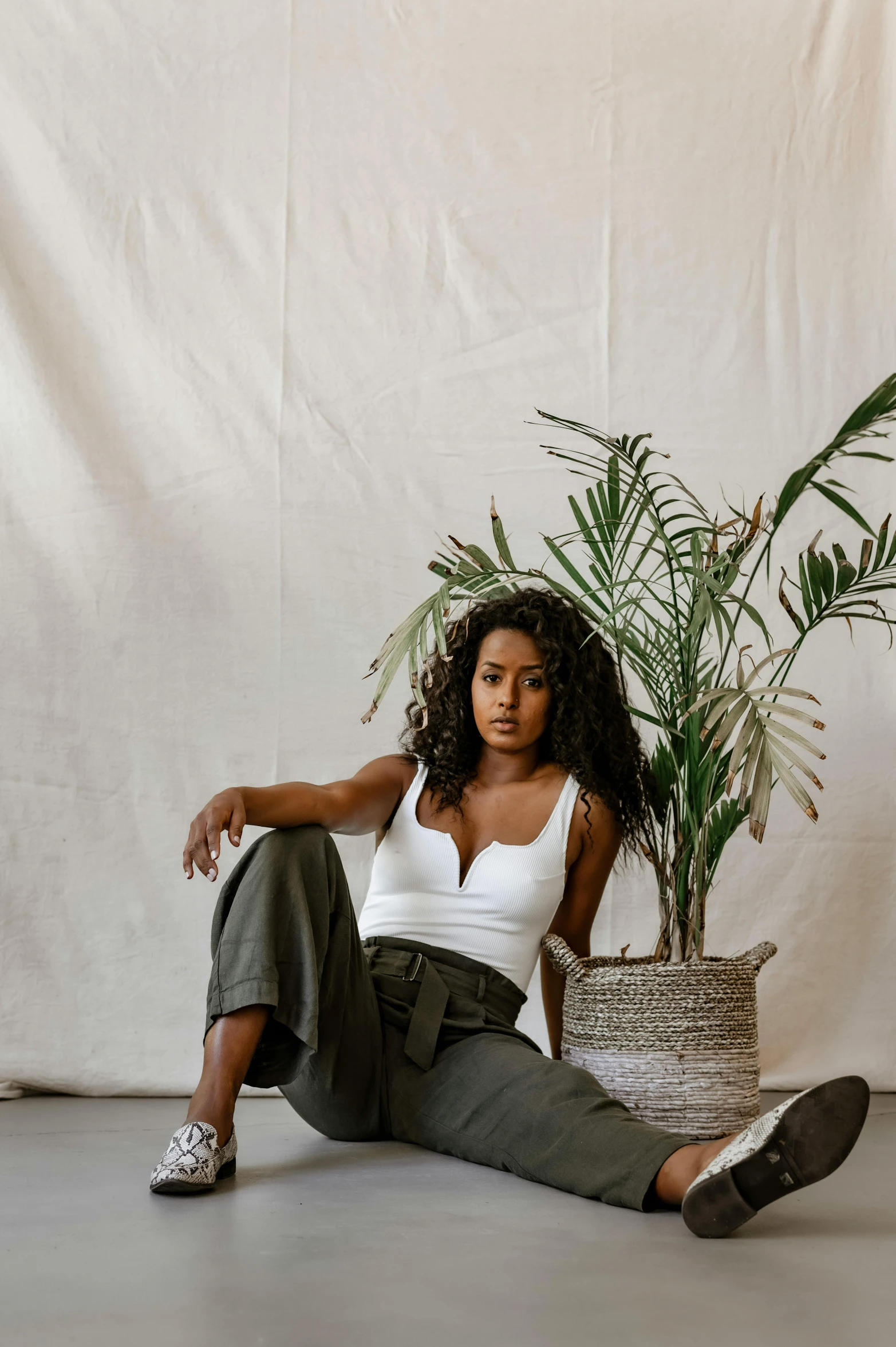 a woman sitting on the floor next to a potted plant, a portrait, inspired by Esaias Boursse, trending on unsplash, she has olive brown skin, olive green slacks, in a white boho style studio, wavy hair spread out