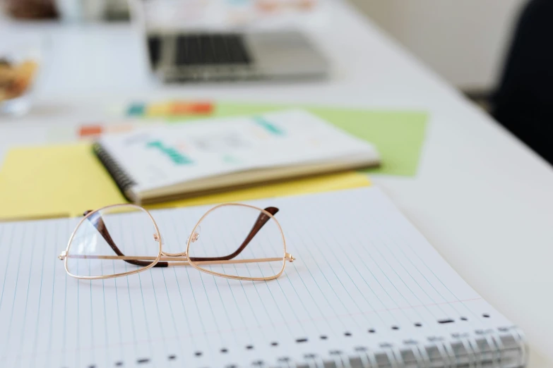 a pair of glasses sitting on top of a notebook, sitting on a desk, on a white table, sat at her desk, educational supplies