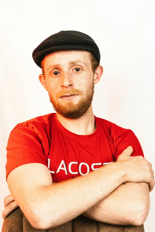 a man sitting on a chair with his arms crossed, an album cover, inspired by Lucas Vorsterman, unsplash, red ascot and a field cap, linus from linustechtips, standup comedian, headshot photograph