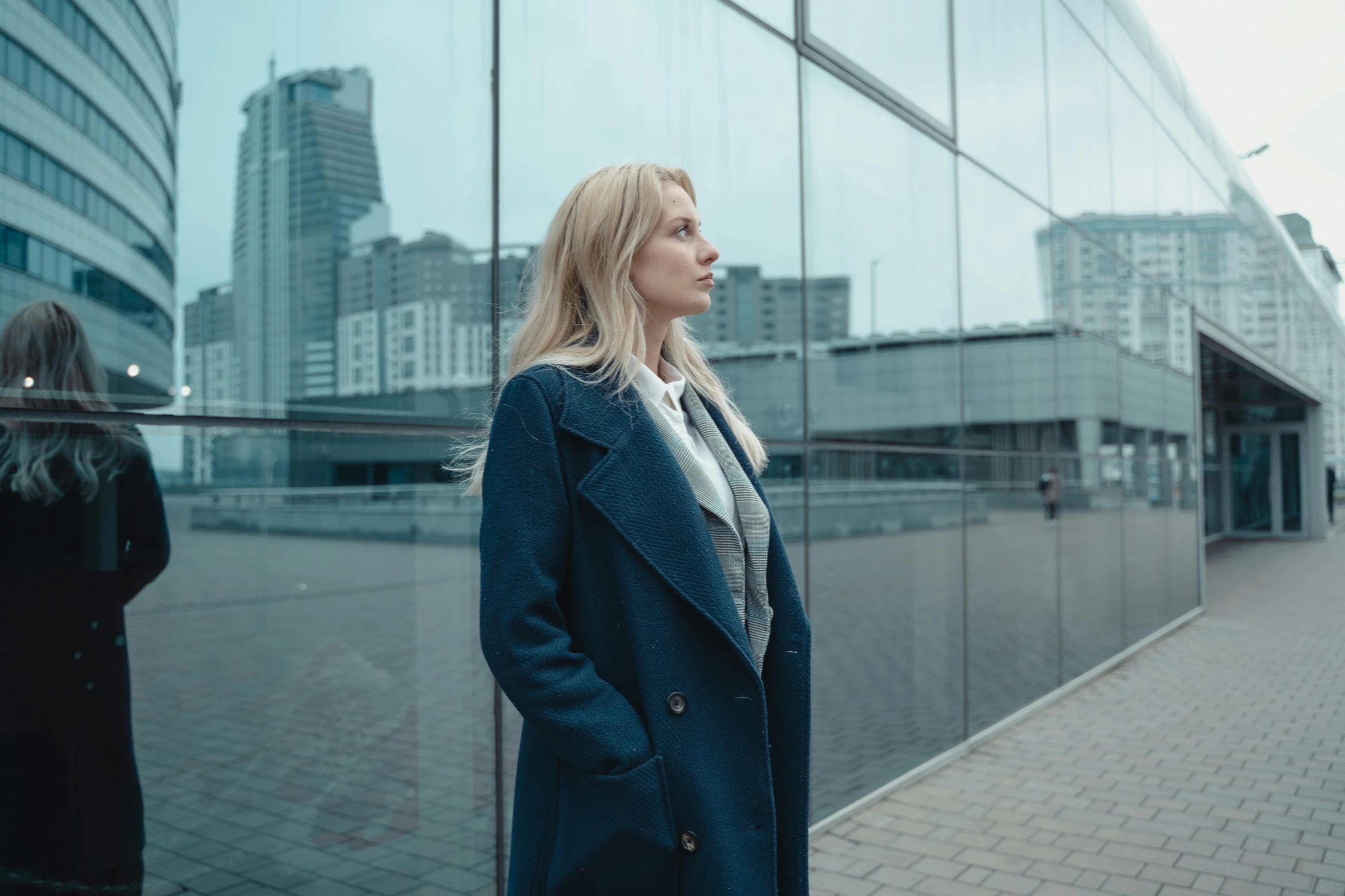 a woman standing in front of a glass building, pexels contest winner, realism, blue coat, young blonde woman, thoughtful ), overcast! cinematic focus