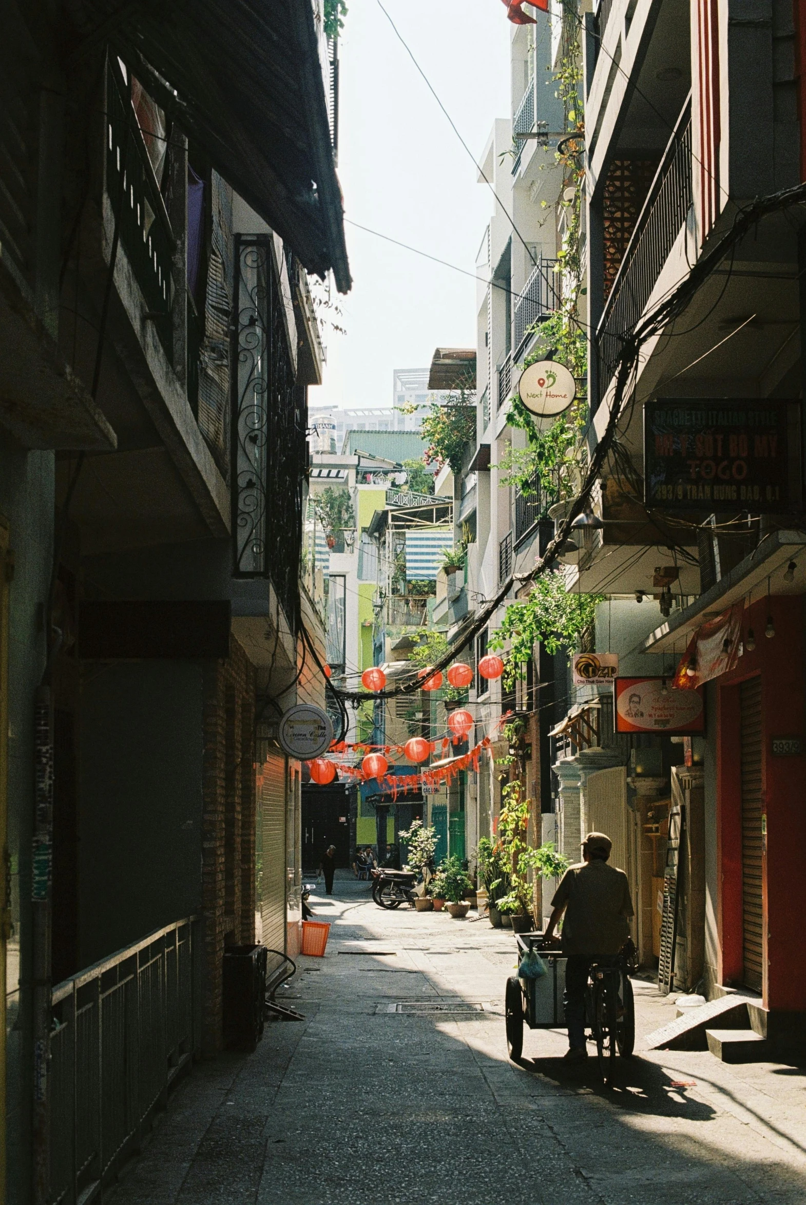 a person riding a motorcycle down a narrow street, by Tan Ting-pho, happening, lots of shops, green alley, on a sunny day, lo fi