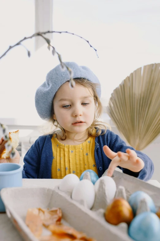 a little girl sitting at a table with eggs, inspired by Elsa Beskow, pexels, blue fedora, spring theme, avatar image, indoor shot