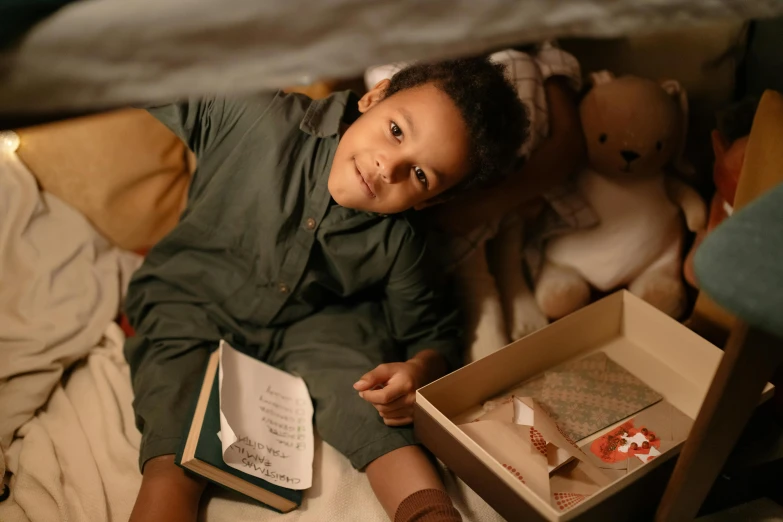 a little girl laying on top of a bed next to a teddy bear, pexels contest winner, happening, he is holding a large book, books cave, ( ( dark skin ) ), looking confident