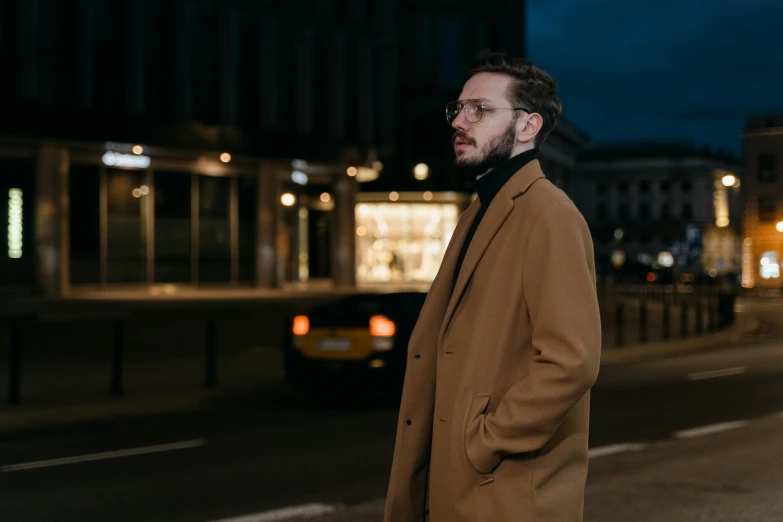 a man standing on a city street at night, an album cover, by Thomas Häfner, pexels contest winner, light brown coat, ginger bearded man with glasses, profile pose, thumbnail