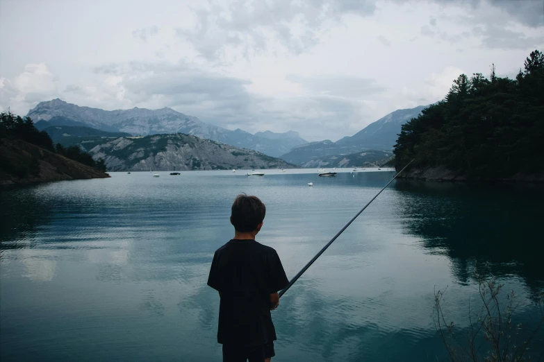 a boy fishing on a lake with mountains in the background, pexels contest winner, shades of blue and grey, childhood friend vibes, traveling in france, young blonde boy fantasy thief