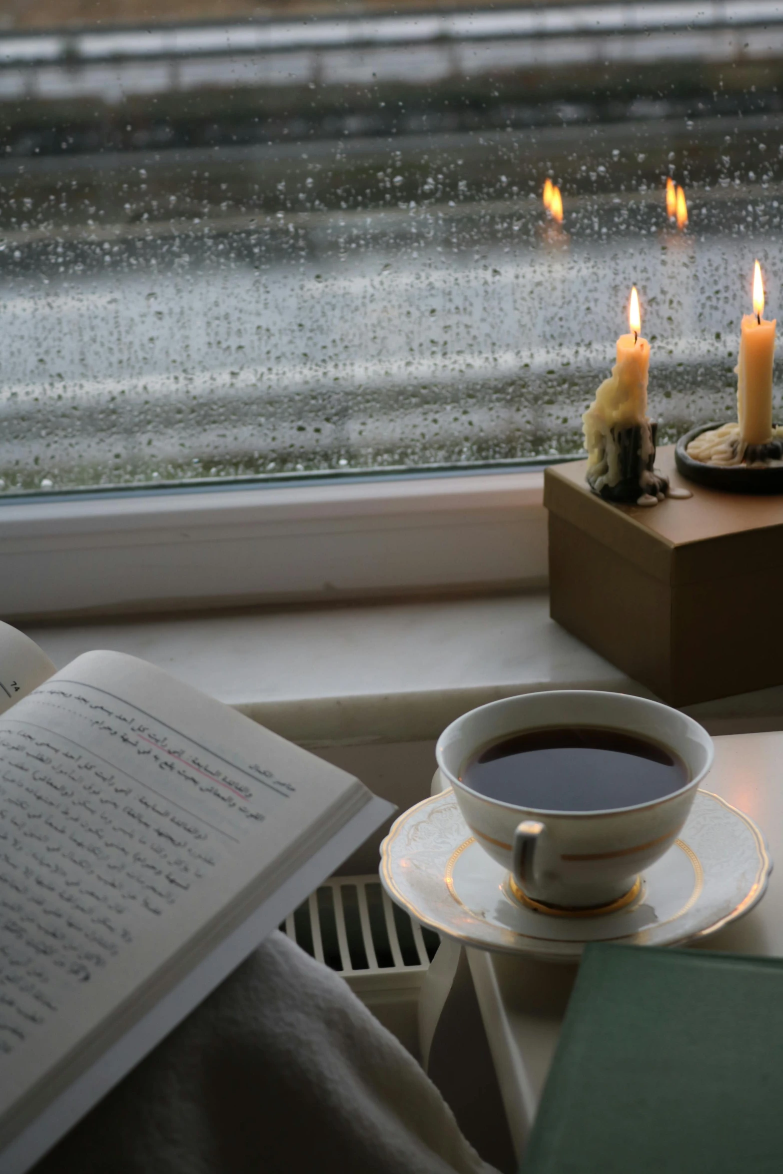 a cup of coffee and a book on a window sill, by Else Alfelt, driving rain, lit candles, during dawn