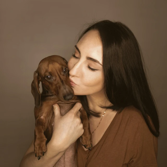 a woman holding a dog in her arms, by Emma Andijewska, pexels contest winner, photorealism, dachshund, cinnamon skin color, studio potrait, kissing together