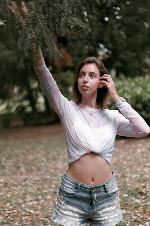 a woman standing under a tree talking on a cell phone, a colorized photo, inspired by Elsa Bleda, pexels contest winner, happening, wearing a sexy cropped top, wearing a light shirt, natural beauty expressive pose, 18 years old
