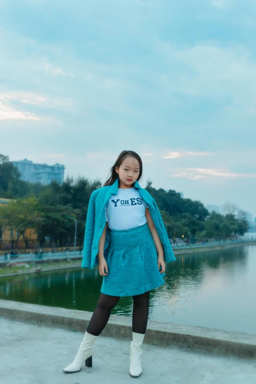 a woman standing in front of a body of water, an album cover, inspired by Kim Tschang Yeul, unsplash, kids, wearing jacket and skirt, lake blue, in a city