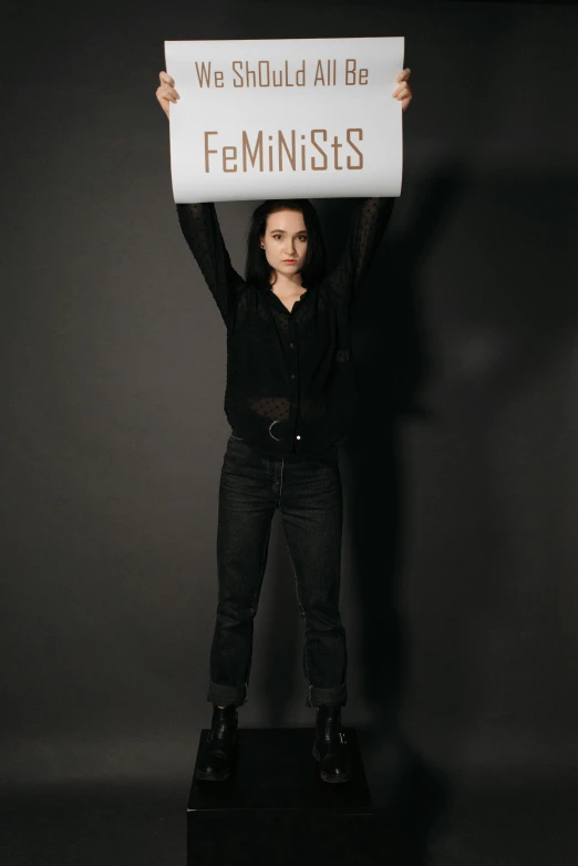 a woman holding a sign that says we should all be feminists, an album cover, by Emma Andijewska, fan bingbing, ( ( ( wearing jeans ) ) ), full body photoshoot, 15081959 21121991 01012000 4k