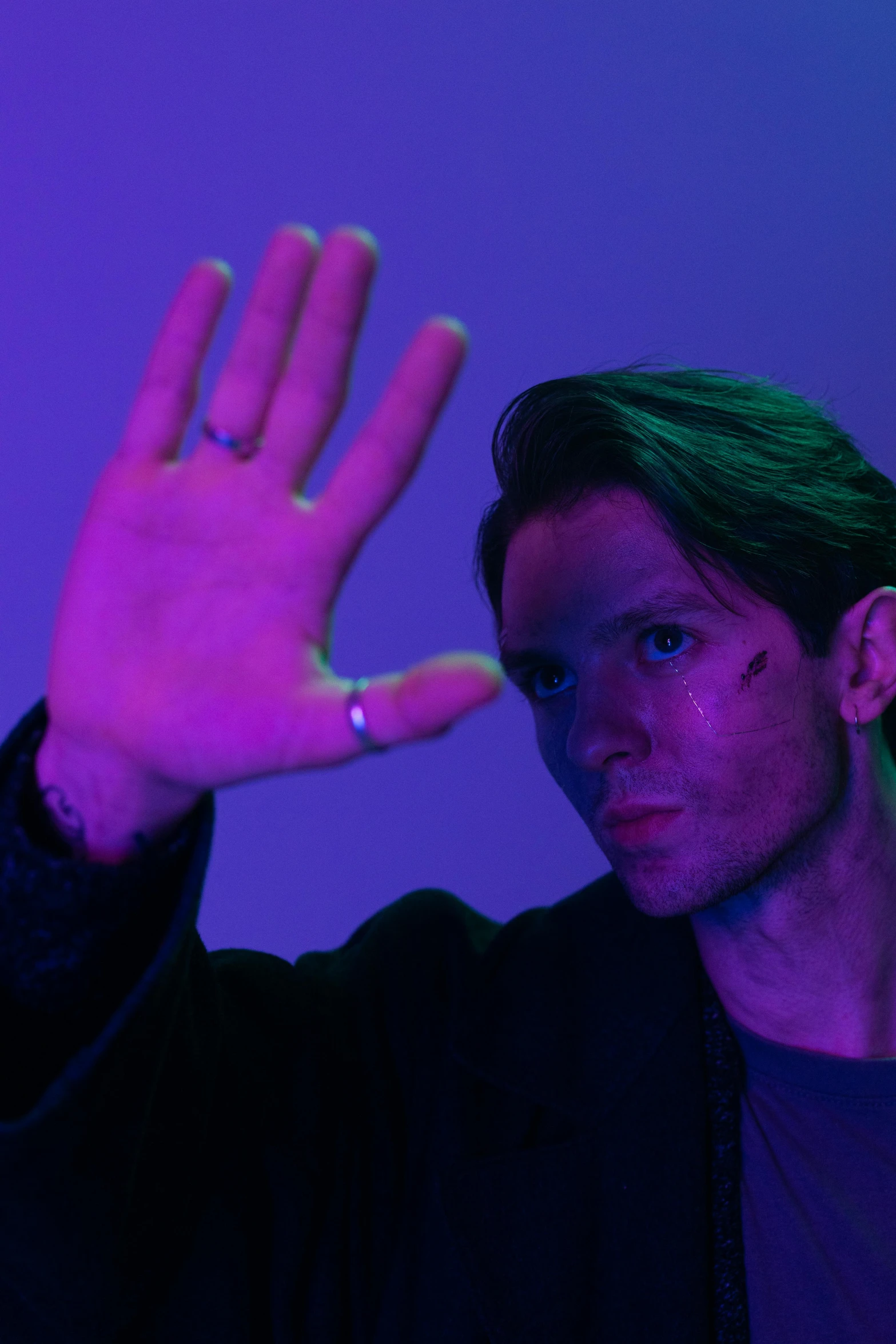 a man holding his hand up in front of his face, an album cover, unsplash, bauhaus, purple light, androgynous male, cheekbones, looking threatening