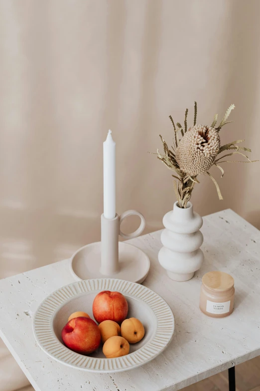 a table topped with a bowl of fruit next to a candle, a still life, unsplash, beige cream natural muted tones, holding a candle holder, product display, tall