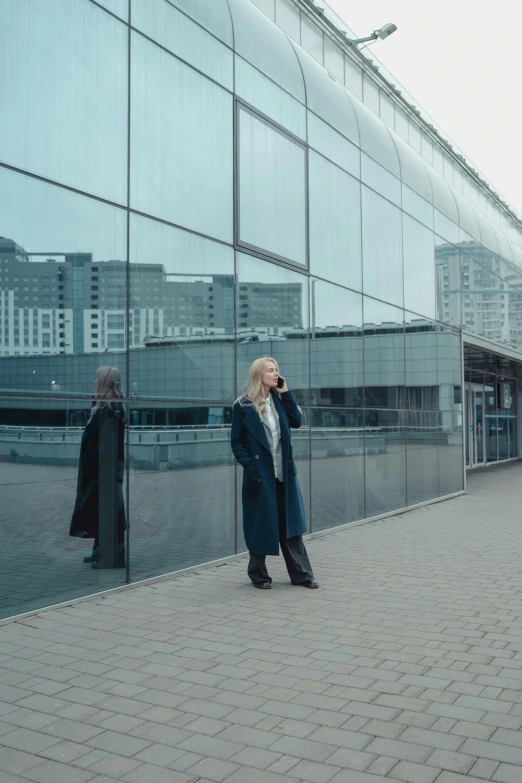 a woman standing on a sidewalk talking on a cell phone, inspired by Elsa Bleda, pexels contest winner, modernism, in moscow centre, glass walls, technological longcoat, standing in an arena