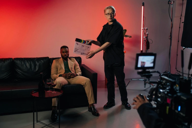 a man standing next to a man sitting on a couch, a picture, by David Donaldson, pexels, performing a music video, shot on arri alexa, mkbhd, jim jarmusch