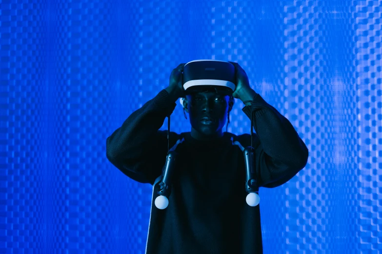 a man wearing a virtual reality headset in front of a blue background, by Adam Marczyński, unsplash, afrofuturism, standing in a dimly lit room, instagram post, blue-black, teamlab