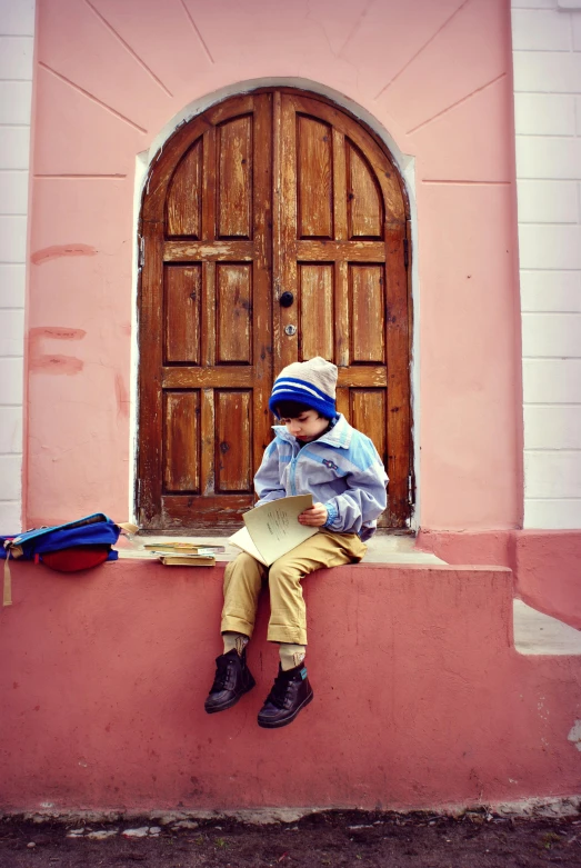 a little boy that is sitting on a wall, pexels contest winner, quito school, holding a book, door, beanie, style of wes anderson