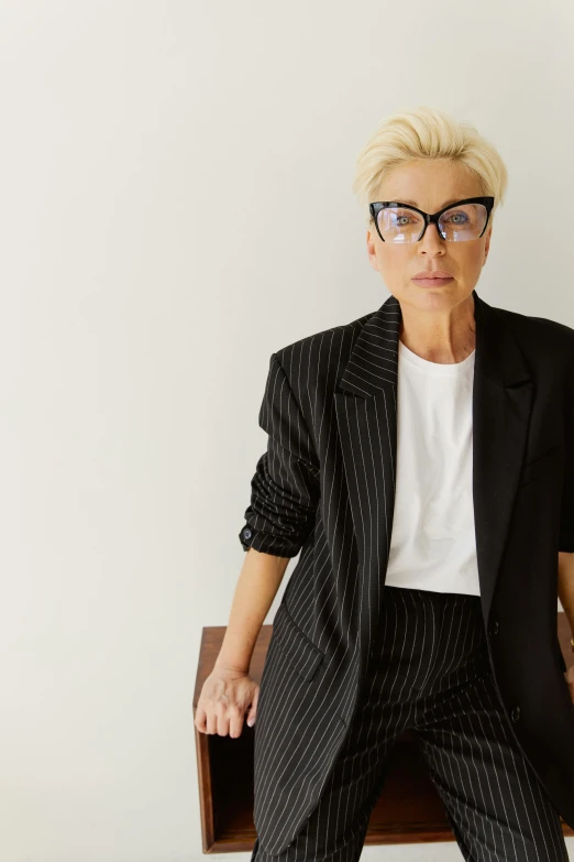 a woman sitting on top of a wooden chair, by Myra Landau, minimalism, in suit with black glasses, 5 5 yo, a blond, androgyny