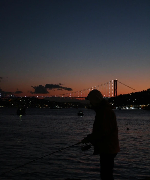 a person fishing in a body of water with a bridge in the background, an album cover, pexels contest winner, hurufiyya, istanbul, dim dusk lighting, low quality footage, sports photo