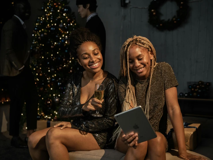 two women sitting on a couch in front of a christmas tree, a portrait, trending on pexels, happening, dark-skinned, using a magical tablet, drinking champagne, people enjoying the show