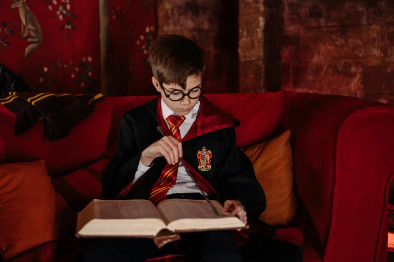 a person sitting on a couch reading a book, a portrait, by Fuller Potter, pexels contest winner, magical school student uniform, hogwarts gryffindor common room, embroidered robes, teenage boy