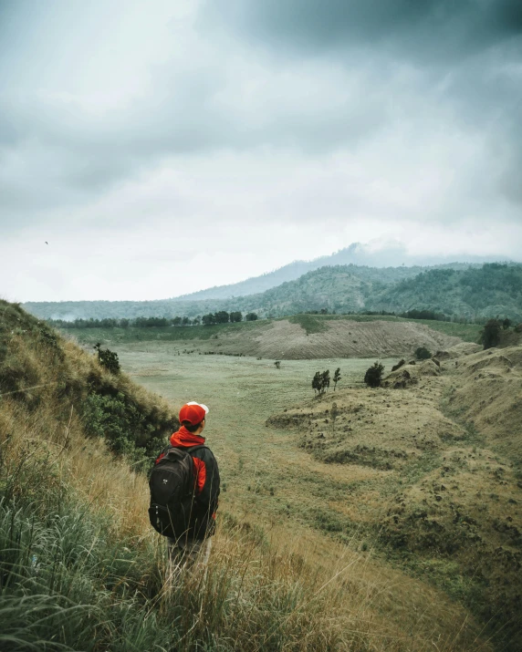 a person walking up a hill on a cloudy day, sumatraism, he has a red hat, instagram post, hunting, valley in the distance