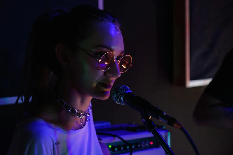 a woman singing into a microphone in front of a keyboard, an album cover, pexels, antipodeans, girl wearing round glasses, low lighting, portrait sophie mudd, live performance