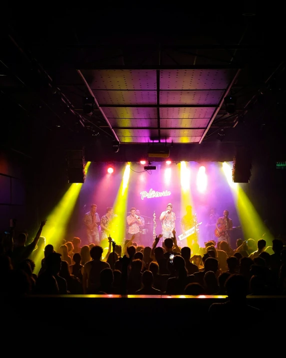 a group of people that are standing in front of a stage, in a nightclub, purple and yellow lighting, big sky, performing