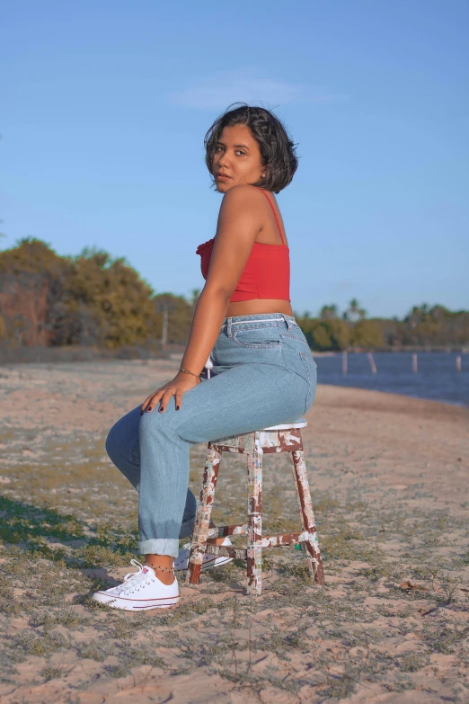 a woman sitting on a stool on a beach, by Lily Delissa Joseph, trending on pexels, happening, red tank top and wide blue pants, ( ( ( wearing jeans ) ) ), side pose, sydney park