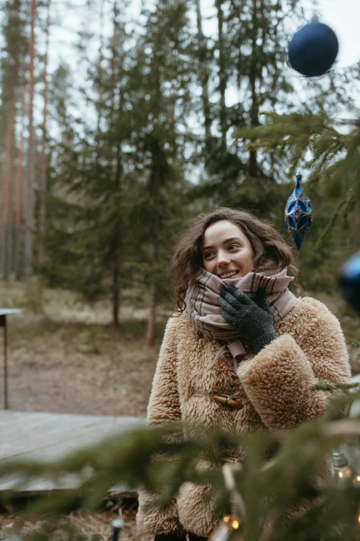 a woman standing in front of a christmas tree, by Adam Marczyński, pexels contest winner, happening, sunny day in the forrest, fur scarf, glamping, dark short curly hair smiling