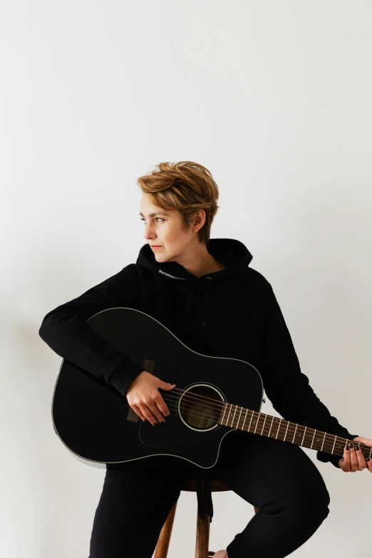 a man sitting on a stool holding a guitar, androgynous face, profile image, in front of white back drop, with short hair