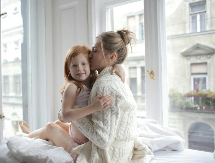 a woman sitting on top of a bed next to a little girl, ginger hair with freckles, hugging each other, looking outside, dressed in a robe