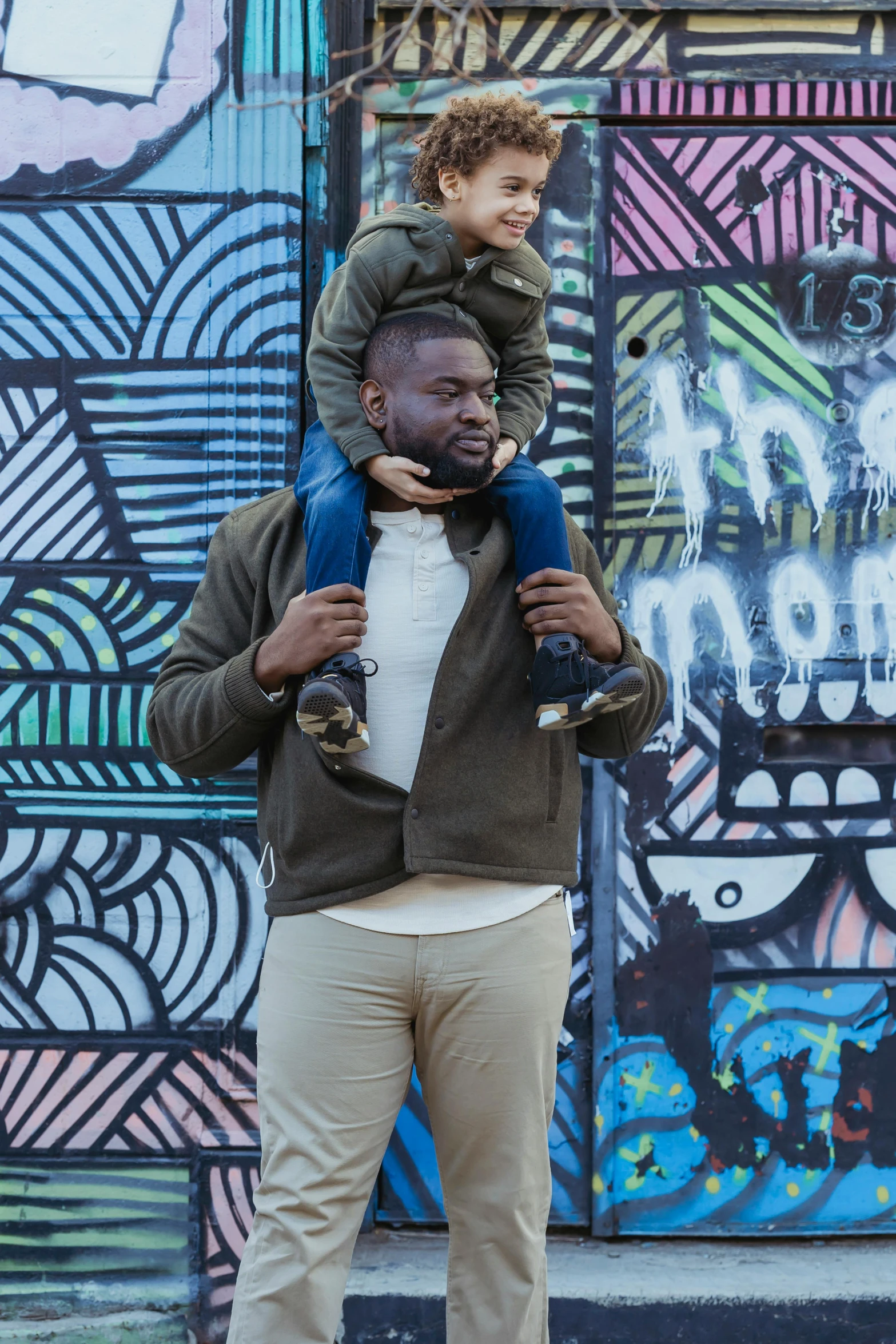 a man carrying a child on his shoulders, by Washington Allston, pexels contest winner, graffiti in background, kevin hart, richly colored, dwell