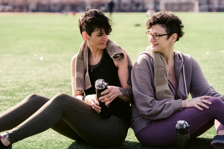 a couple of women sitting on top of a lush green field, a portrait, by Nina Hamnett, pexels, drink milkshakes together, wearing fitness gear, non-binary, brunettes