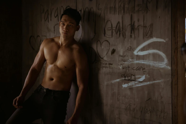 a shirtless man leaning against a wall with graffiti on it, inspired by Ryan Yee, standing in a dimly lit room, [ theatrical ], damien tran, lachlan bailey